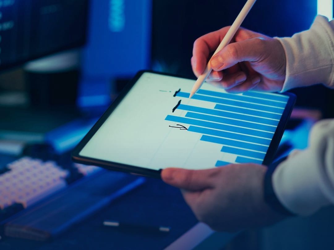 Hands holding a tablet with a blue bar chart and a stylus, analysing business data